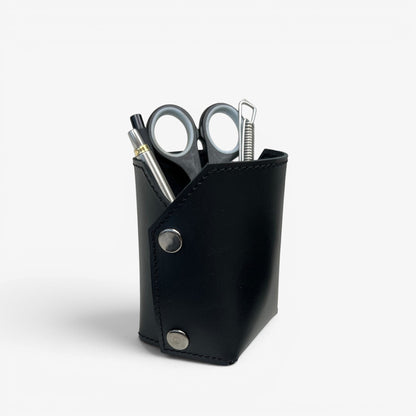 Pache black leather pen and brush holder with silver snap closures, holding pens and scissors on a desk.