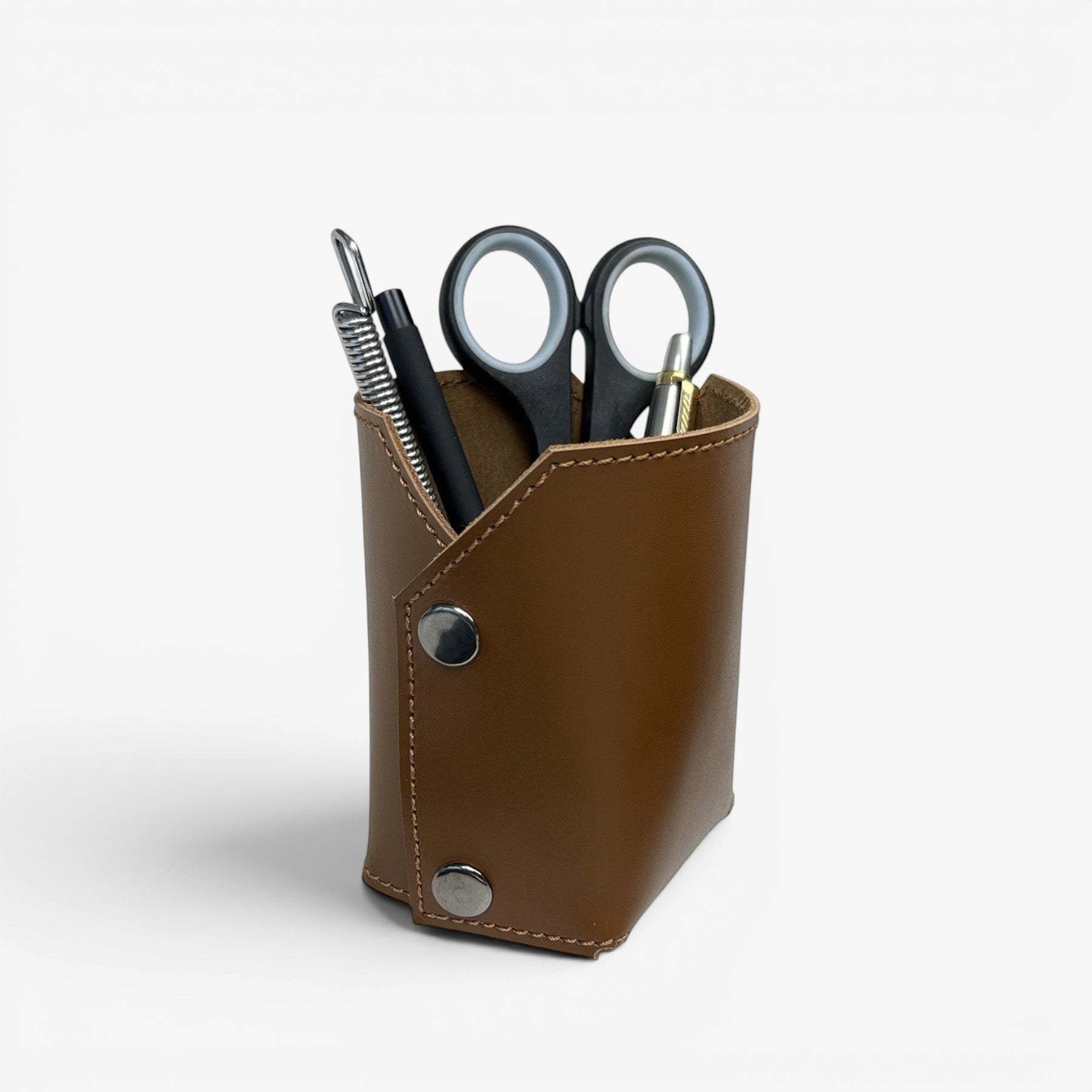 Pache brown leather desk organizer holding pens, scissors, and a plant in a white pot. Minimalist design.