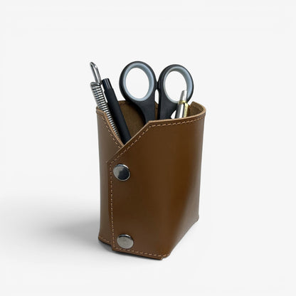 Pache brown leather desk organizer holding pens, scissors, and a plant in a white pot. Minimalist design.
