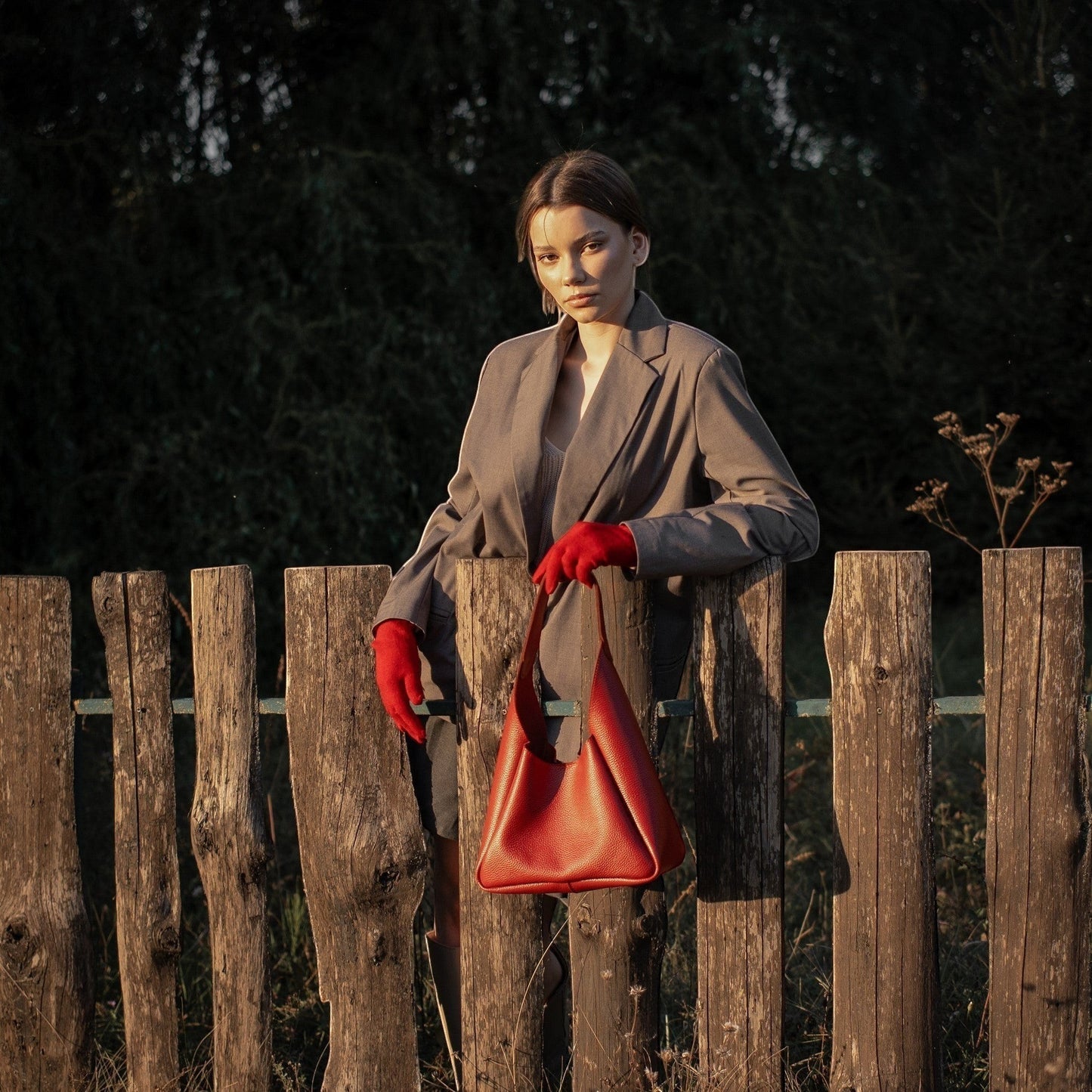 Pache Torba hobo bag in red pebbled leather, held by a woman wearing a grey blazer and red gloves, leaning on a wooden fence.